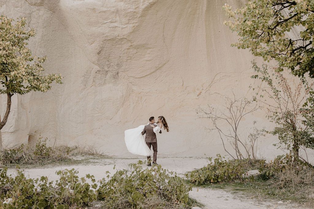 Groom lifting bride in white gown near white rock formation surrounded by lush green trees.