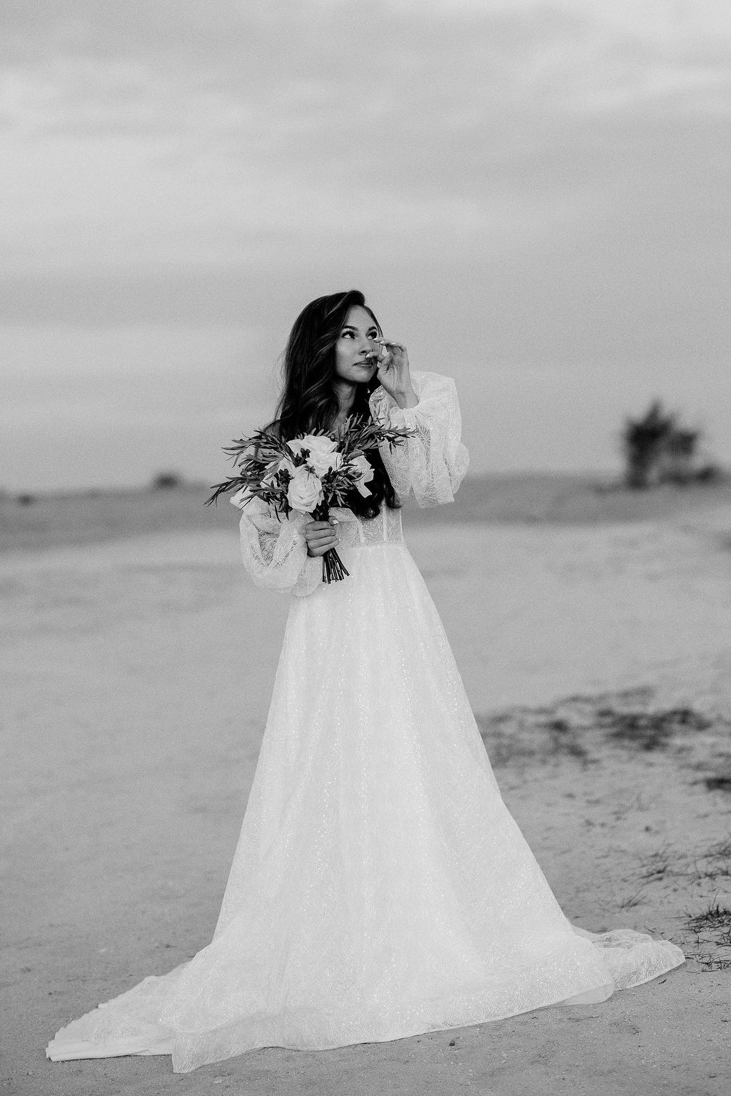 Black and white photo of a bride in a flowing white gown holding a bouquet, standing on a sandy beach under a cloudy sky.