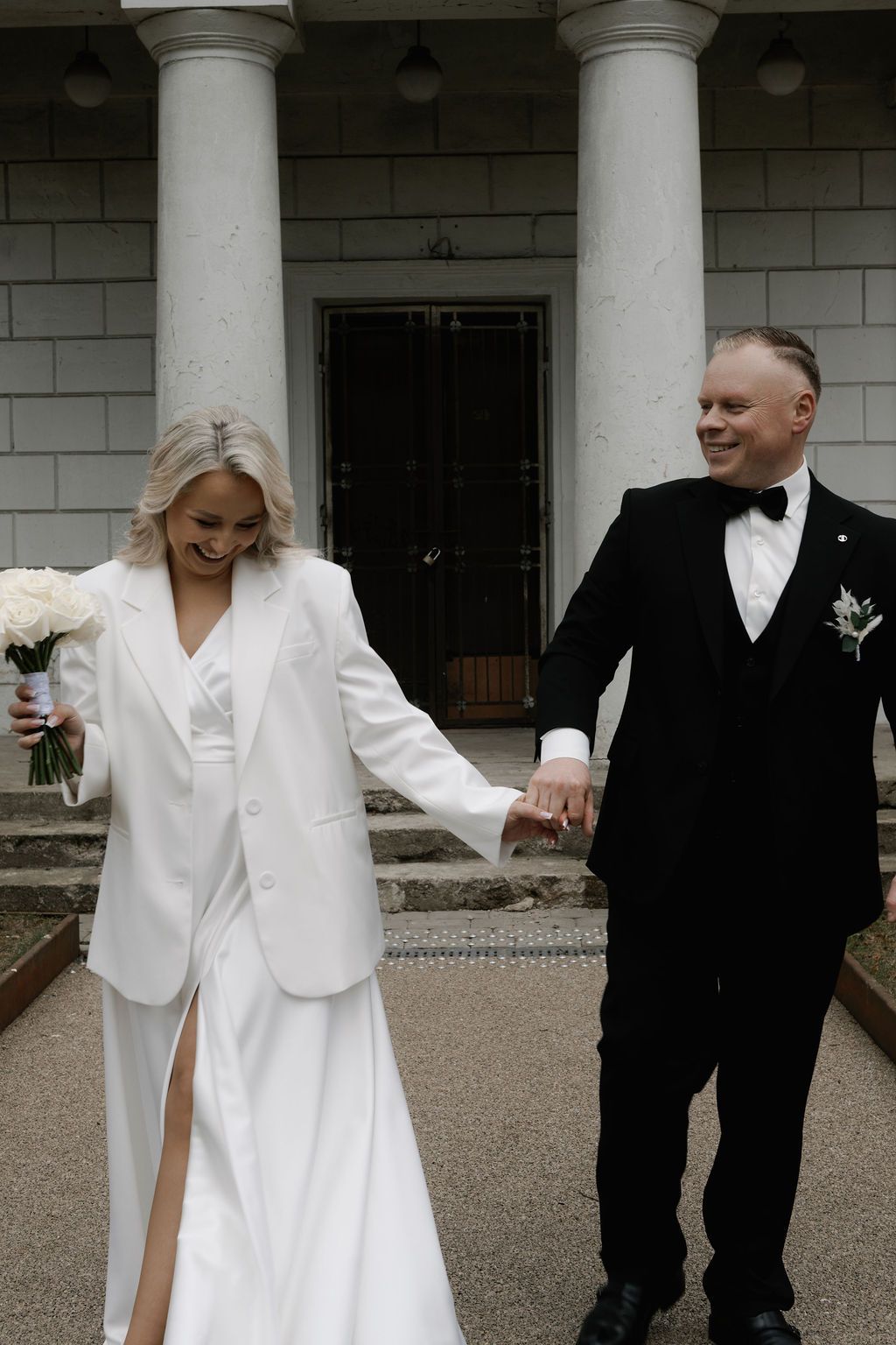 Happy couple in wedding attire descend steps of columned venue, bride in white blazer holding bouquet, groom in black tux.