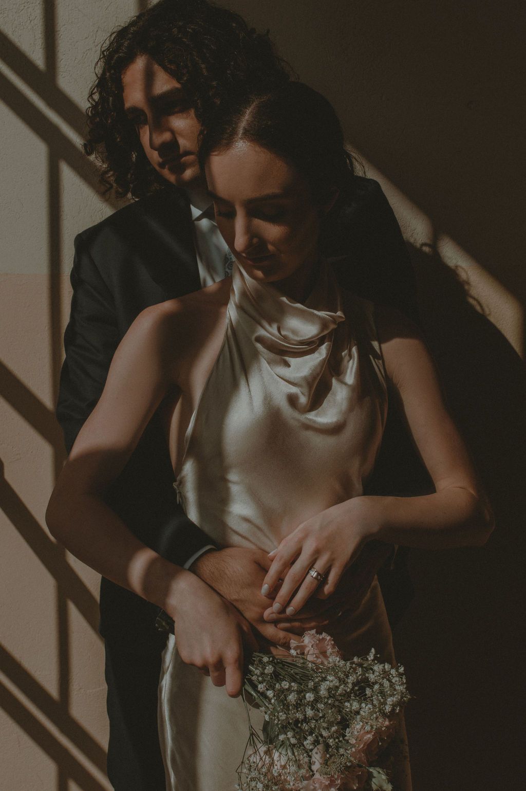 A bride in a silk slip dress holds baby's breath flowers while her groom in a dark suit embraces her from behind in soft light.