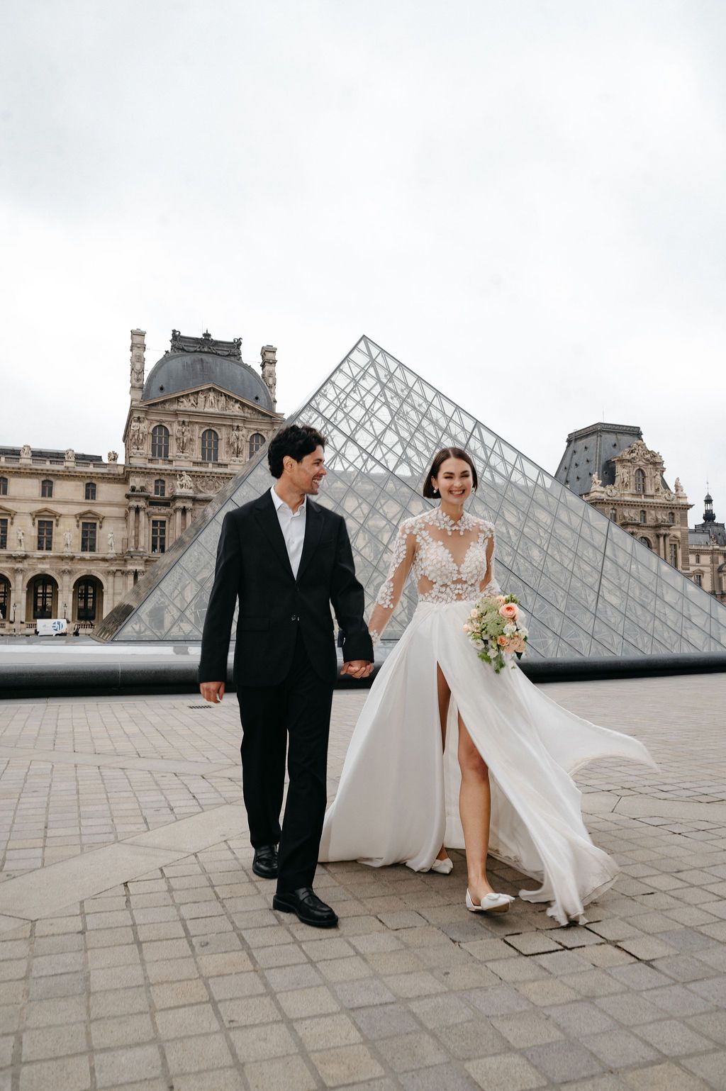 A couple in wedding attire holds hands in front of the Louvre Pyramid in Paris, France.