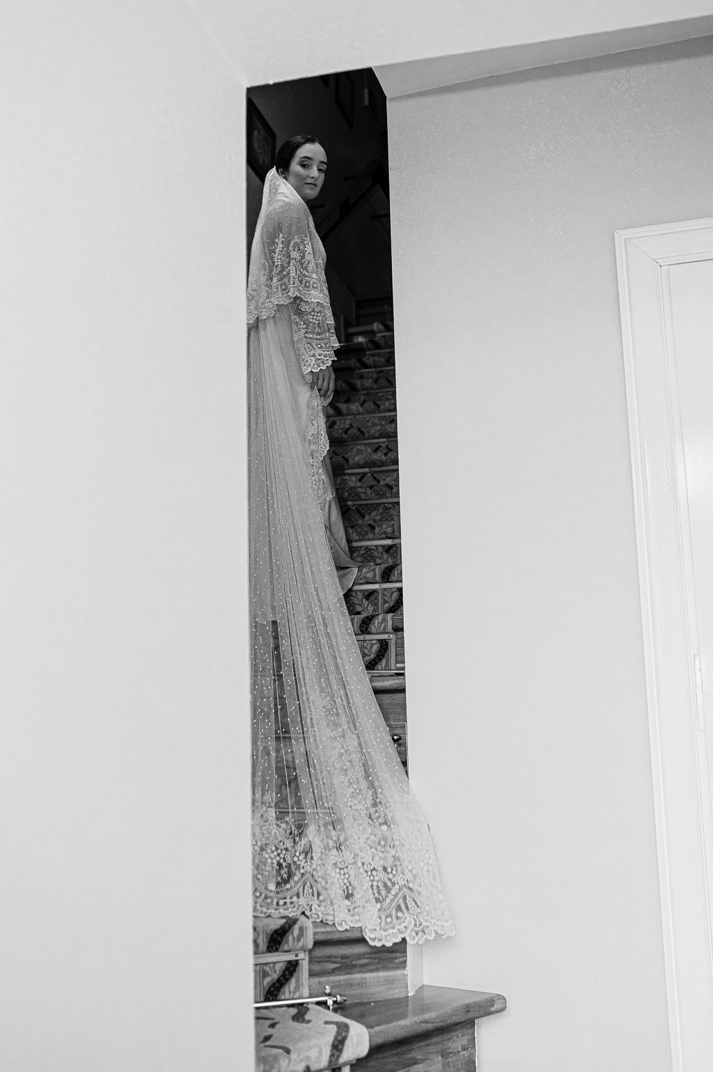 Black and white photo of bride in lace gown and veil standing on staircase.