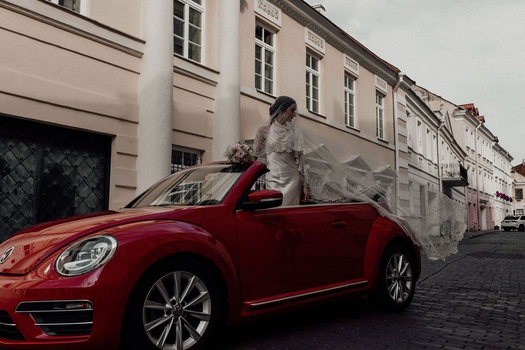 Bride in white wedding dress posing beside a red Volkswagen Beetle convertible on a cobblestone European street.