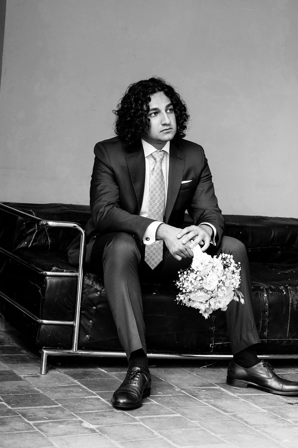Groom in dark suit sits on leather sofa holding bouquet in elegant black and white wedding portrait.