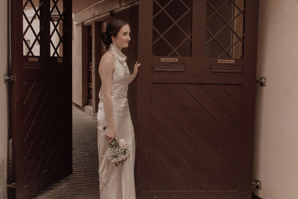 Elegant bride in sleek white gown holding a bouquet, standing by vintage wooden doors in a European setting.