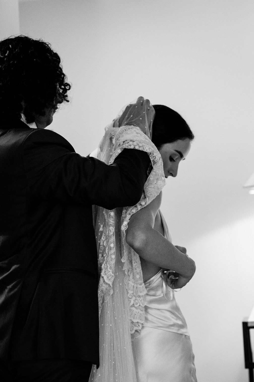 Black and white photo of a person helping a bride with her lace veil and wedding dress buttons before the ceremony.
