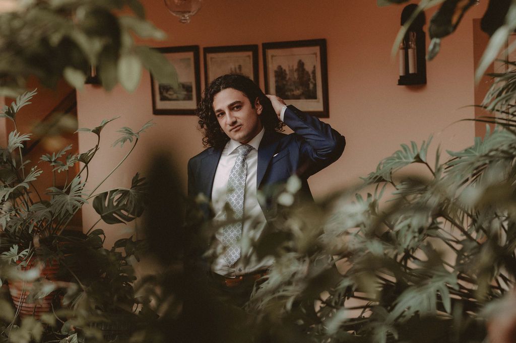 Man in blue suit and tie posing among lush green plants in a warmly lit room with framed art on walls.