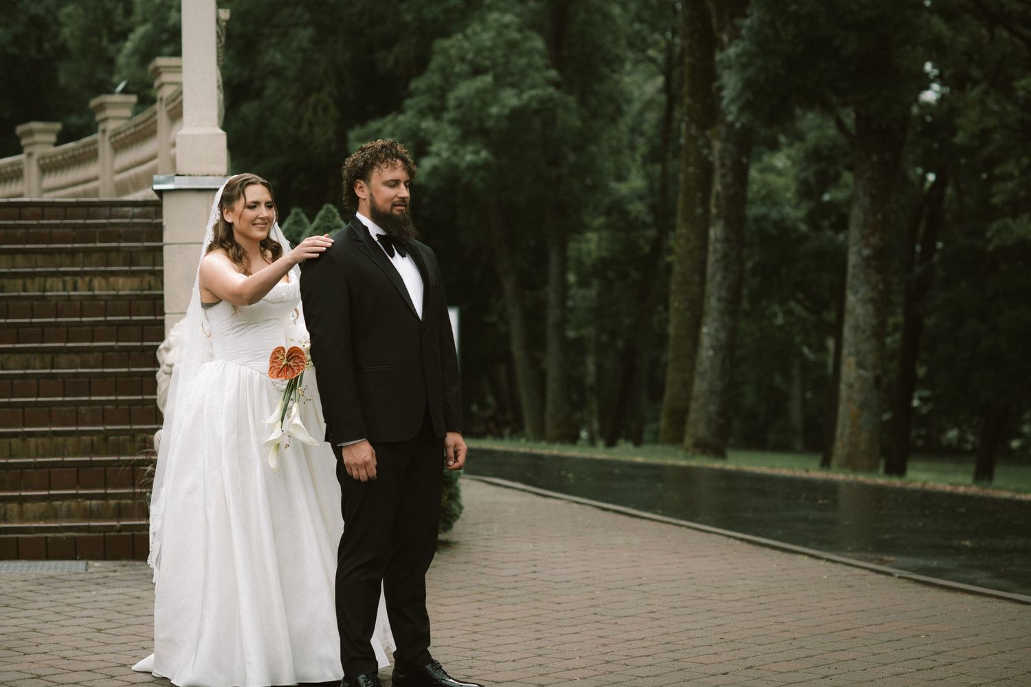 Bride and groom posing outdoors in a lush green park, wearing wedding attire with a white gown and black tuxedo.
