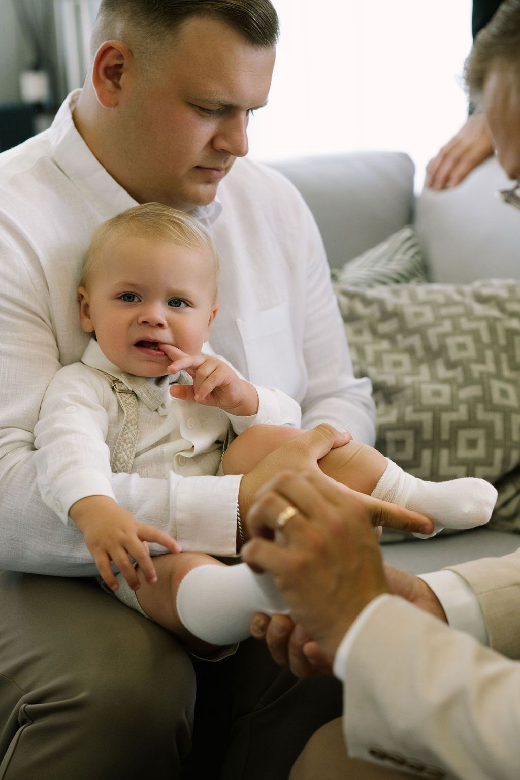 Father in white shirt holds baby in white outfit on sofa while another person touches infant's foot.