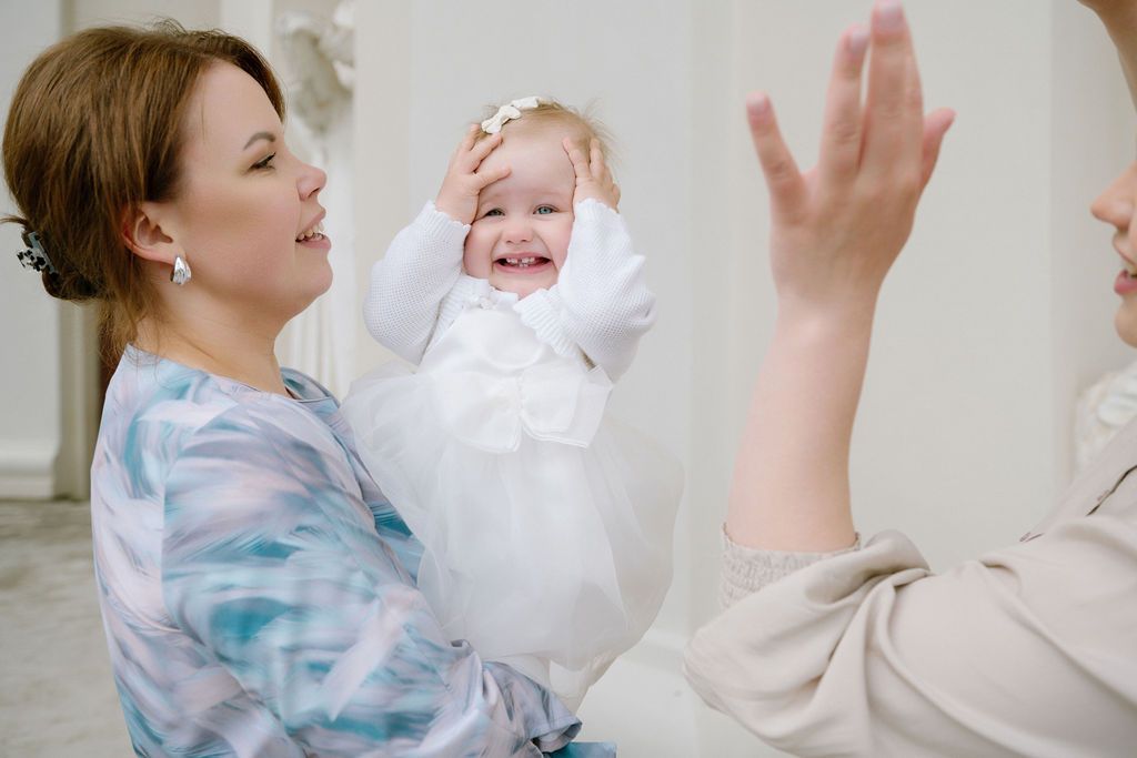 A smiling woman holds a laughing baby in a white dress at a christening ceremony.