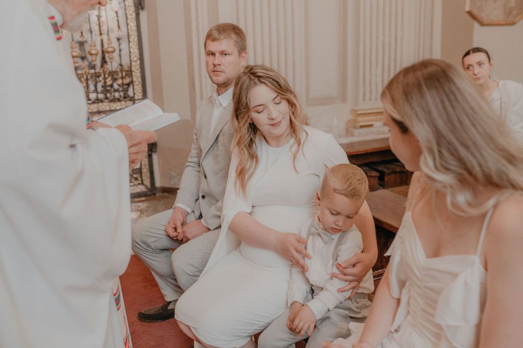 Family gathered in church for a baptism ceremony, with a pregnant mother holding a toddler beside a priest.