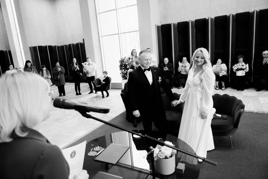 Black and white wedding ceremony photo with bride in white gown and groom in tuxedo before officiant.