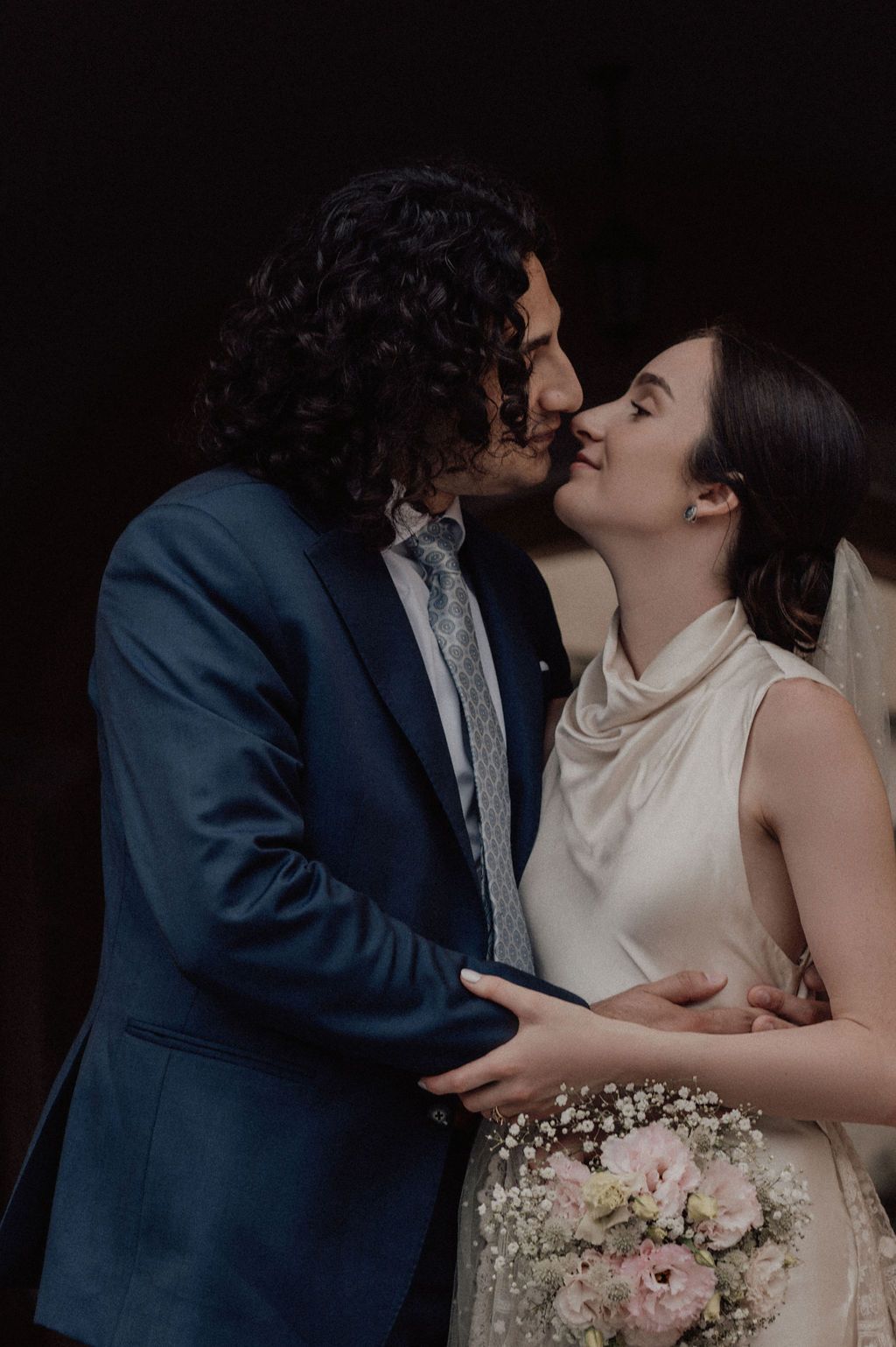 A couple in wedding attire share a romantic kiss, the groom in a blue suit and bride holding a bouquet of pink flowers.
