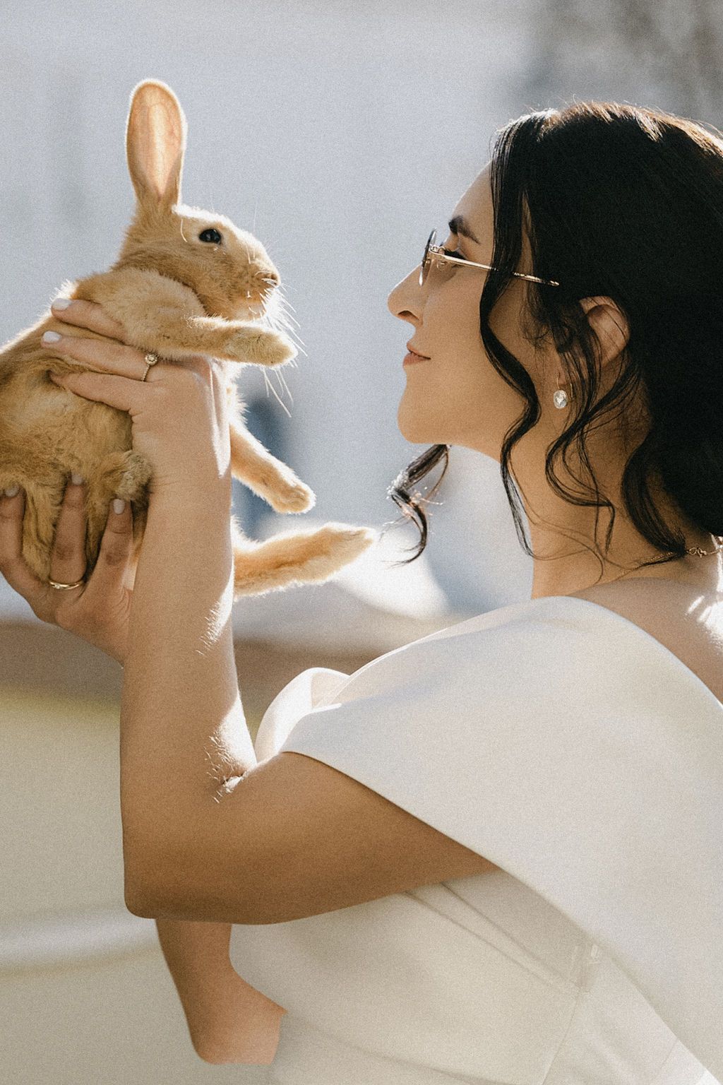 Elegant bride in white dress holding a cute tan rabbit up to her face in soft natural light.