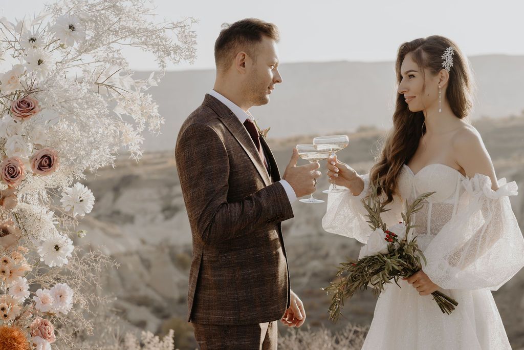 Couple in wedding attire toasting with champagne glasses outdoors, groom in plaid suit, bride in off-shoulder gown holding bouquet.