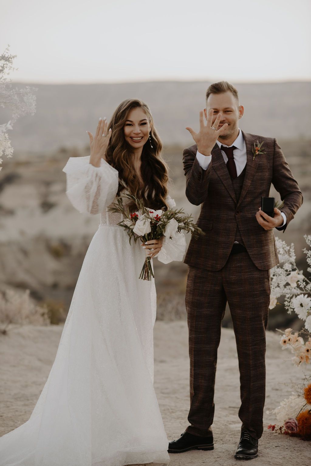 Bride and groom showing wedding rings outdoors in desert landscape, bride holding bouquet in white gown, groom in brown plaid suit.