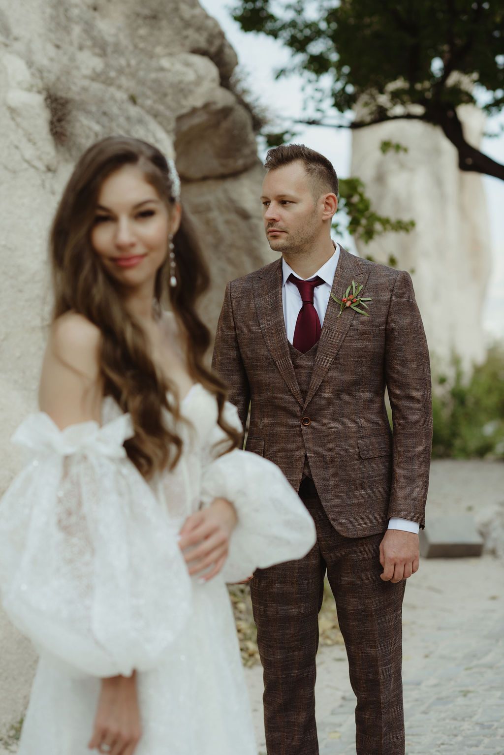 Bride in white lace dress with groom in brown plaid suit and burgundy tie at outdoor wedding.