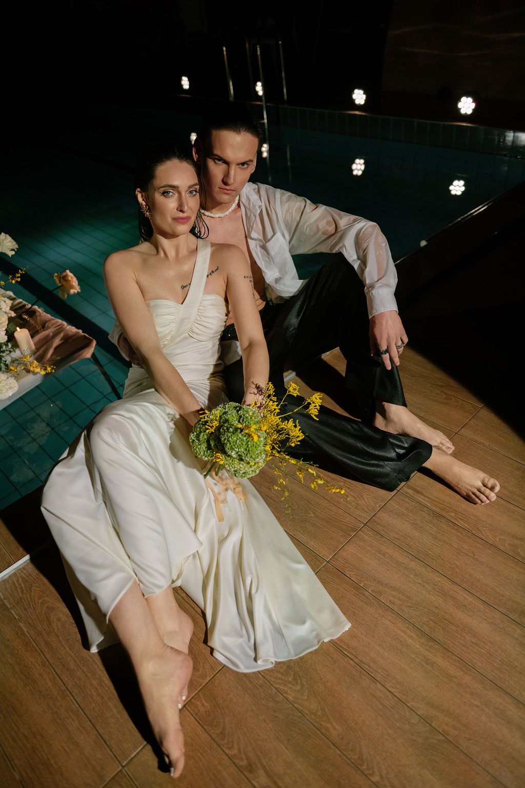 Elegant bride in white gown holds yellow bouquet beside groom in white shirt near poolside lights.