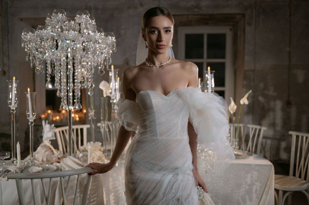 Elegant bride in a strapless white tulle wedding gown stands near crystal candelabras in a romantic reception venue.