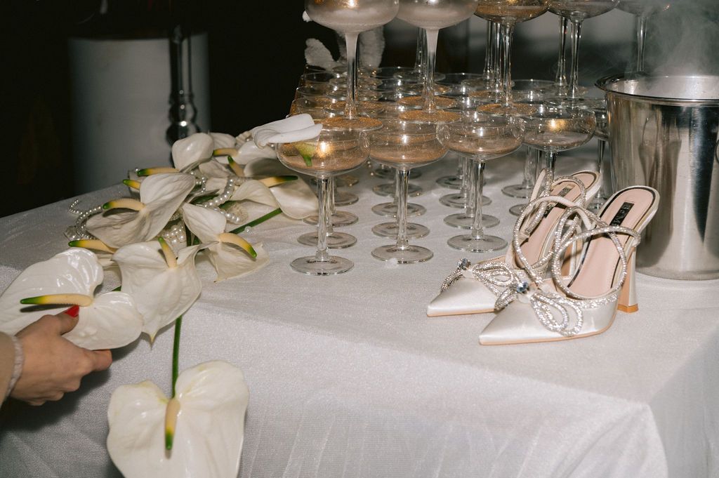 Elegant white heels and white orchids displayed on a white tablecloth beside champagne glasses at a wedding reception.