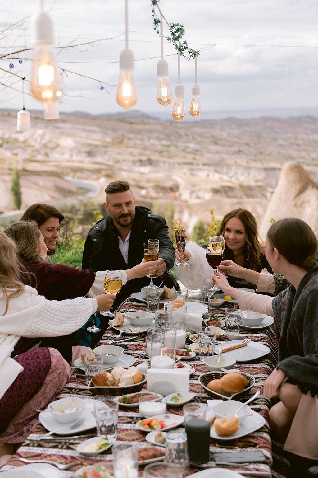 Friends toasting with wine glasses at an outdoor dining table with scenic Cappadocia landscape and string lights.