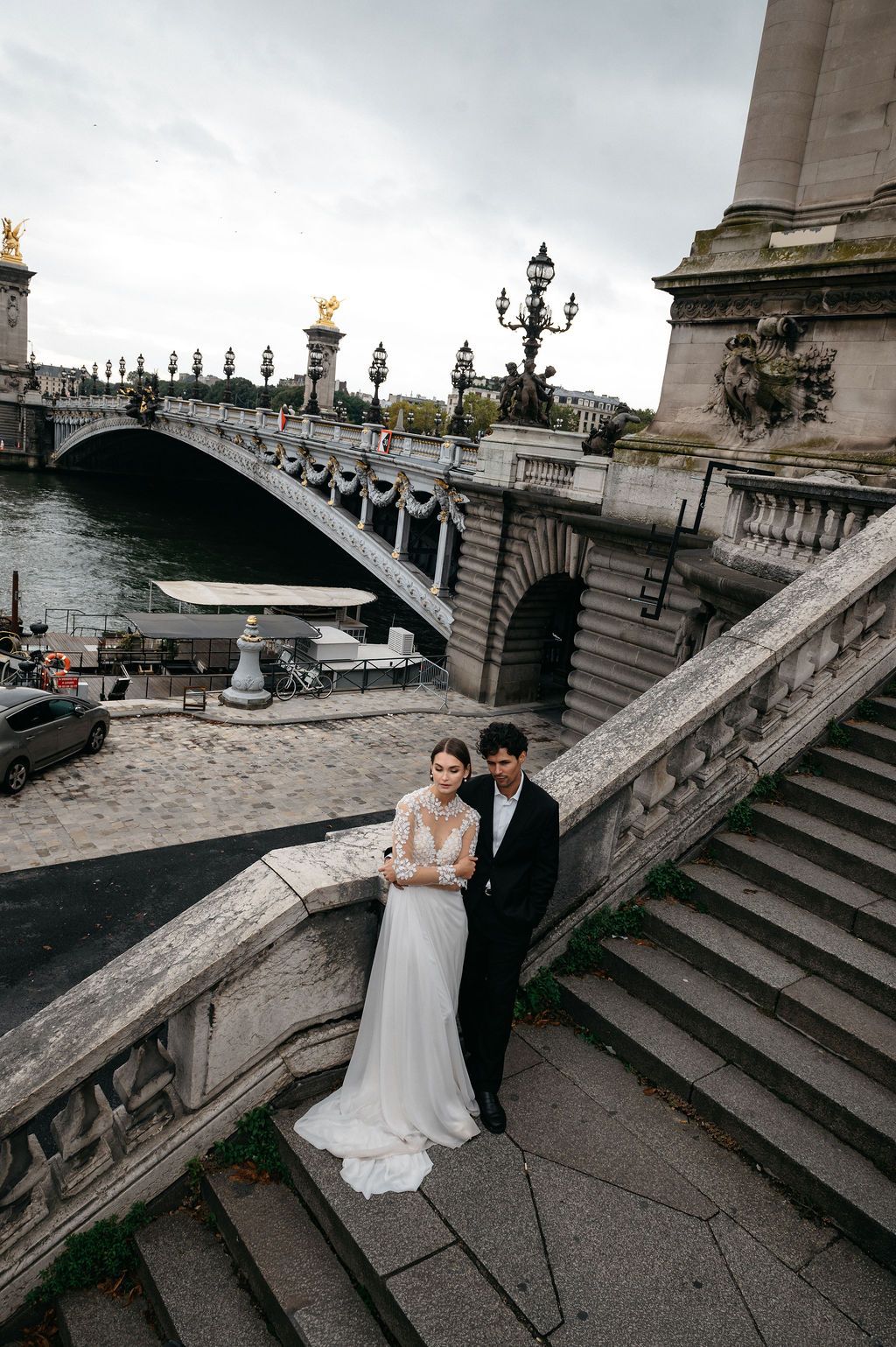 Bride and groom posing on ornate stone steps near Pont Alexandre III bridge in Paris on their wedding day.
