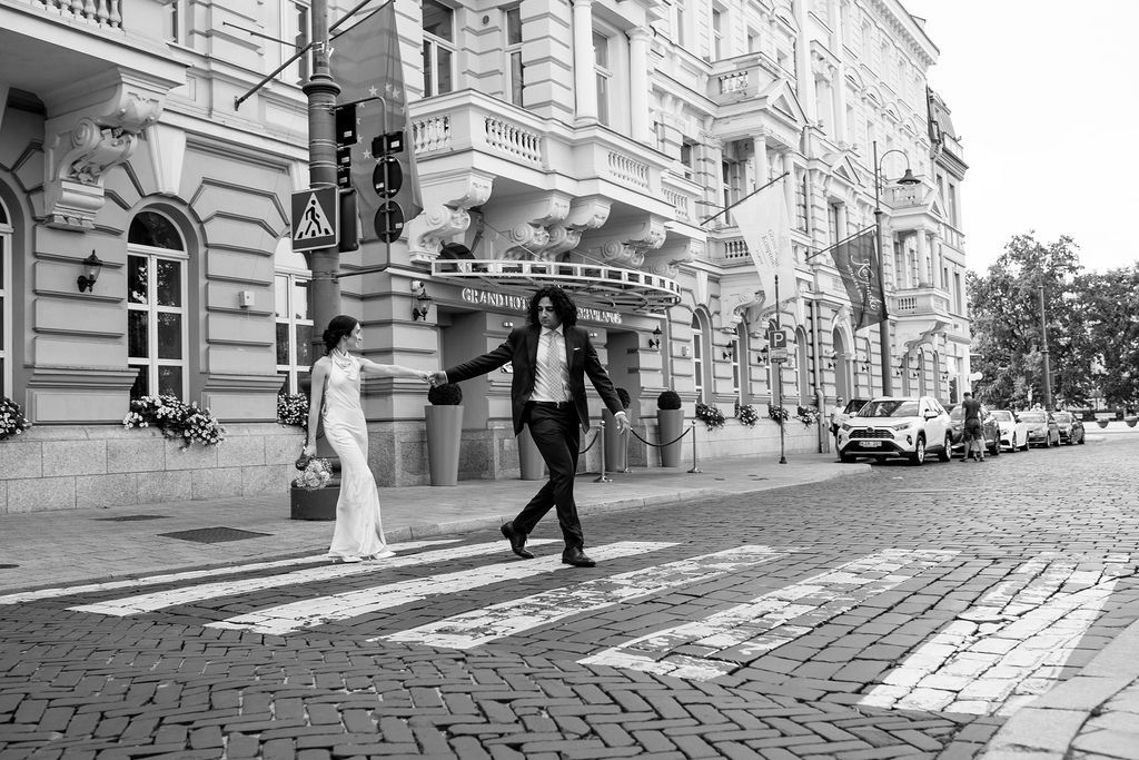 Black and white photo of bride and groom holding hands crossing a zebra crossing on a European city street.