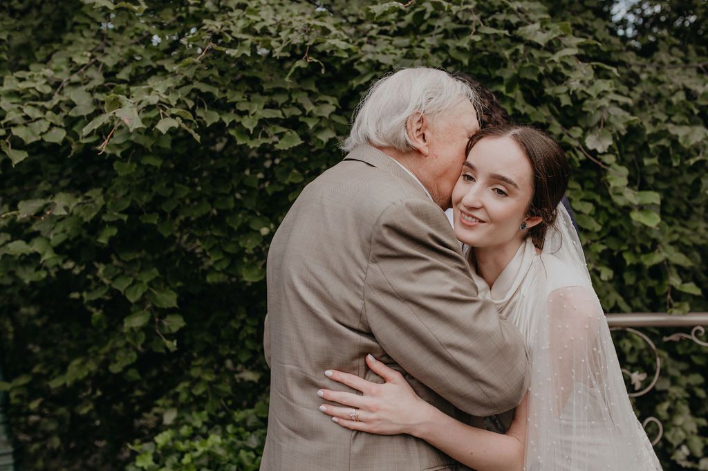 Elderly grandfather tenderly kissing bride on cheek during emotional wedding day embrace outdoors.