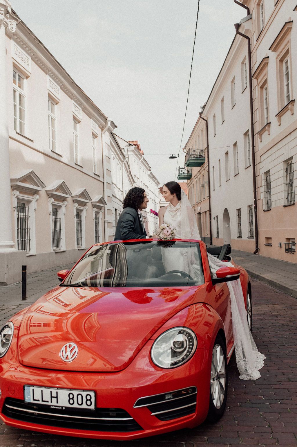 Bride and groom standing in red VW Beetle convertible on a European cobblestone street.