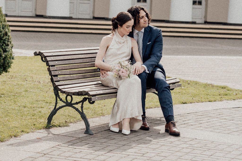 A bride and groom sit closely together on a wooden bench outdoors, she holds a bouquet in a white dress.