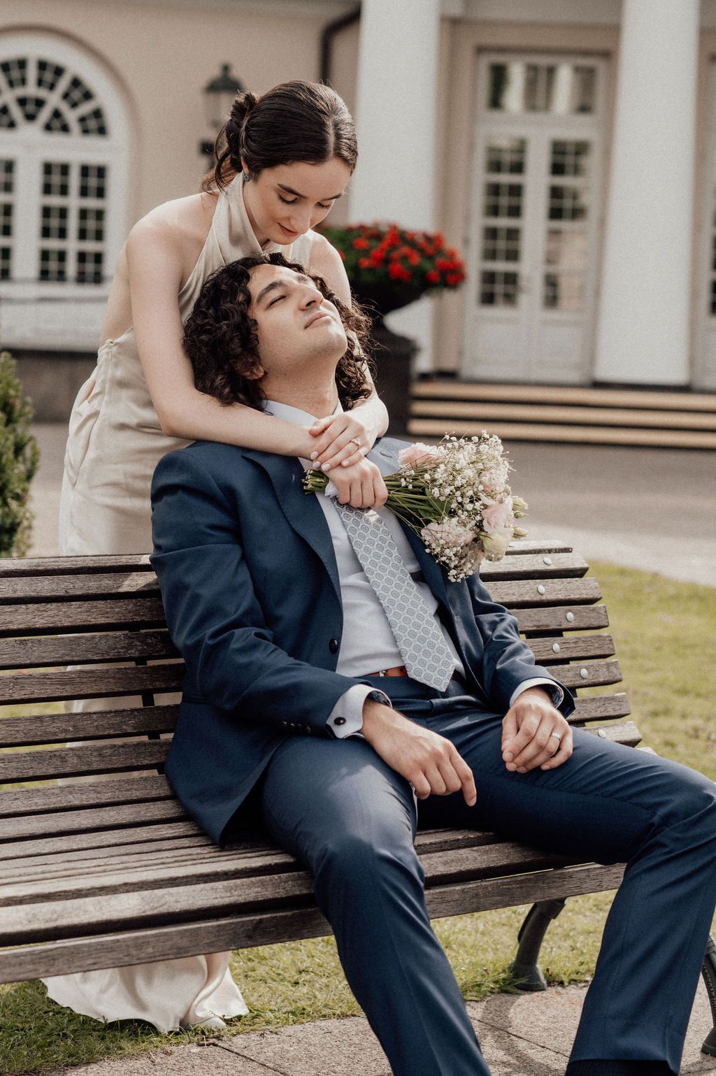 A groom in a navy suit sits on a bench as his bride in a white dress leans over him holding a bouquet.