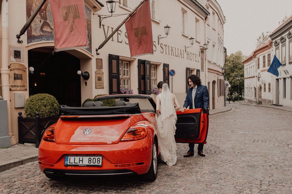 Bride and groom beside a red VW Beetle convertible on a cobblestone street outside a historic European hotel.