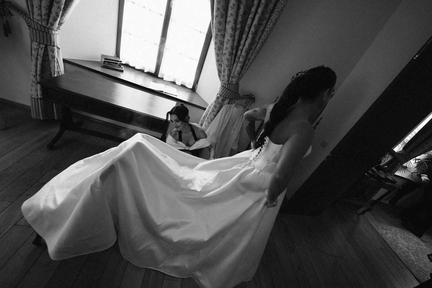 Black and white photo of a bride in a flowing white gown standing in a sunlit room as a young girl sits near a window.