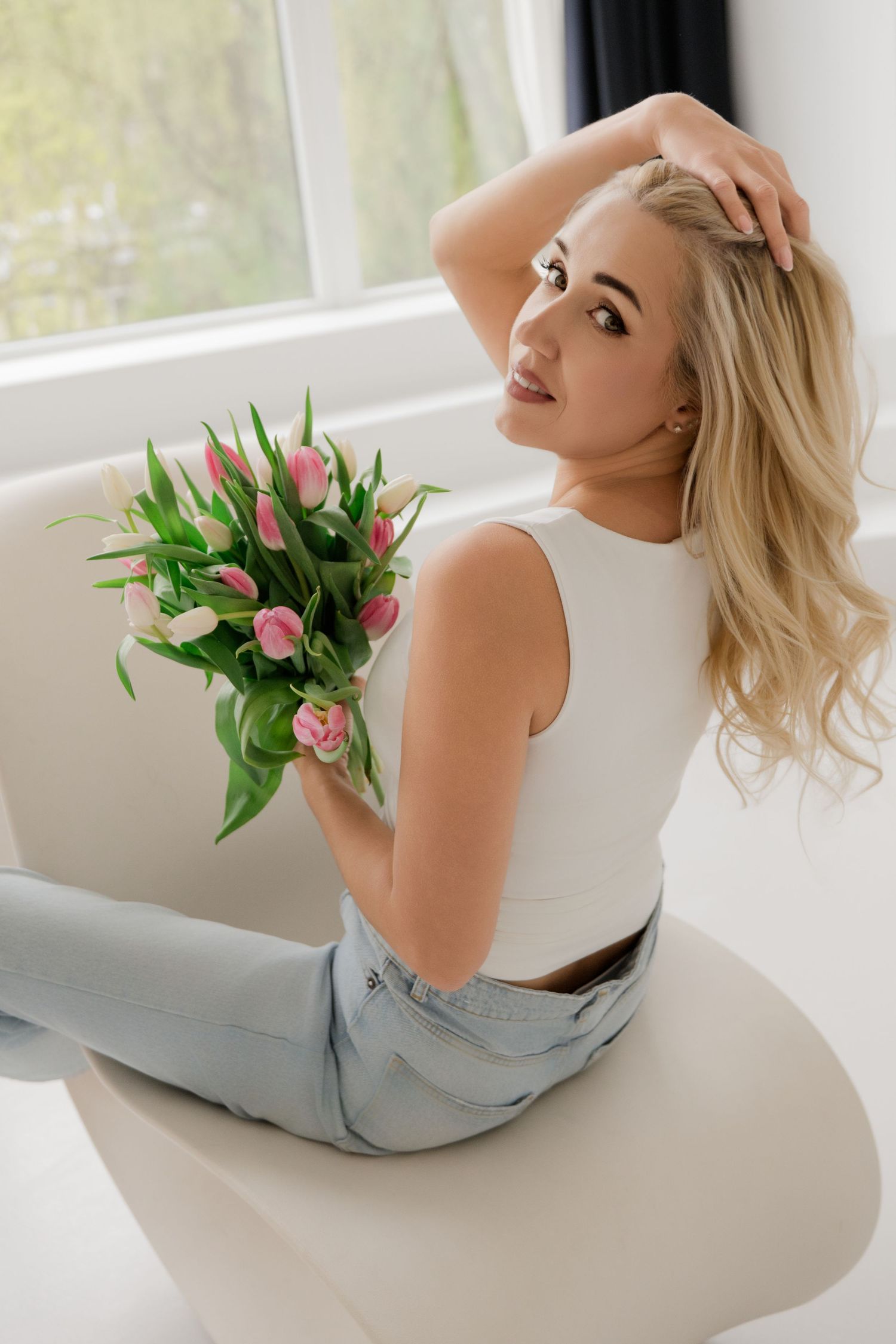 Blonde woman in white crop top and jeans sitting on chair holding pink tulips bouquet near bright window.
