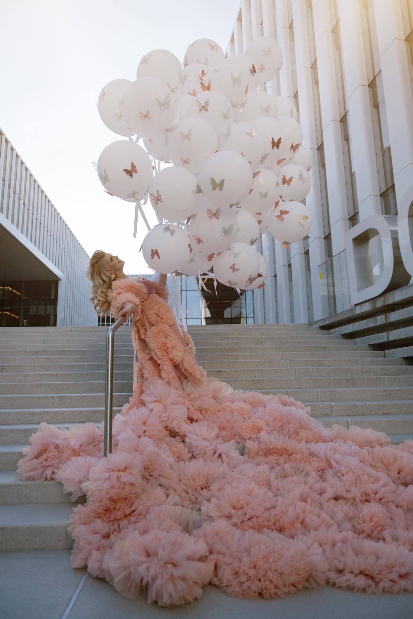 Woman in pink ruffled gown holding white butterfly balloons on grand steps of modern building.