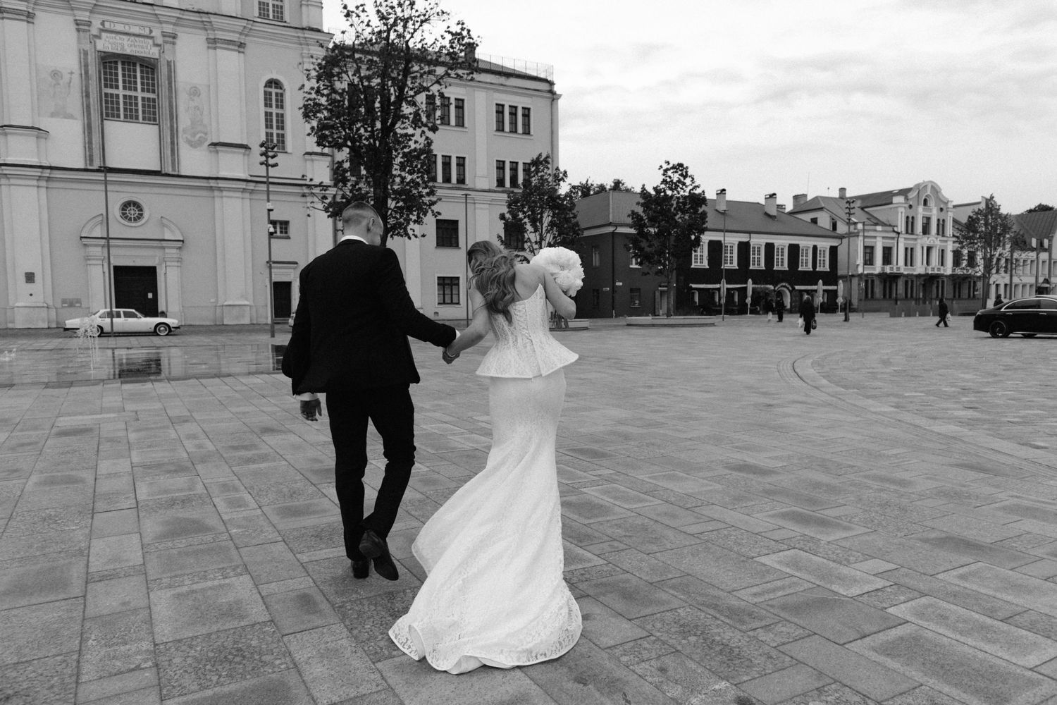 Black and white photo of a bride and groom walking hand in hand across a cobblestone plaza near historic buildings.