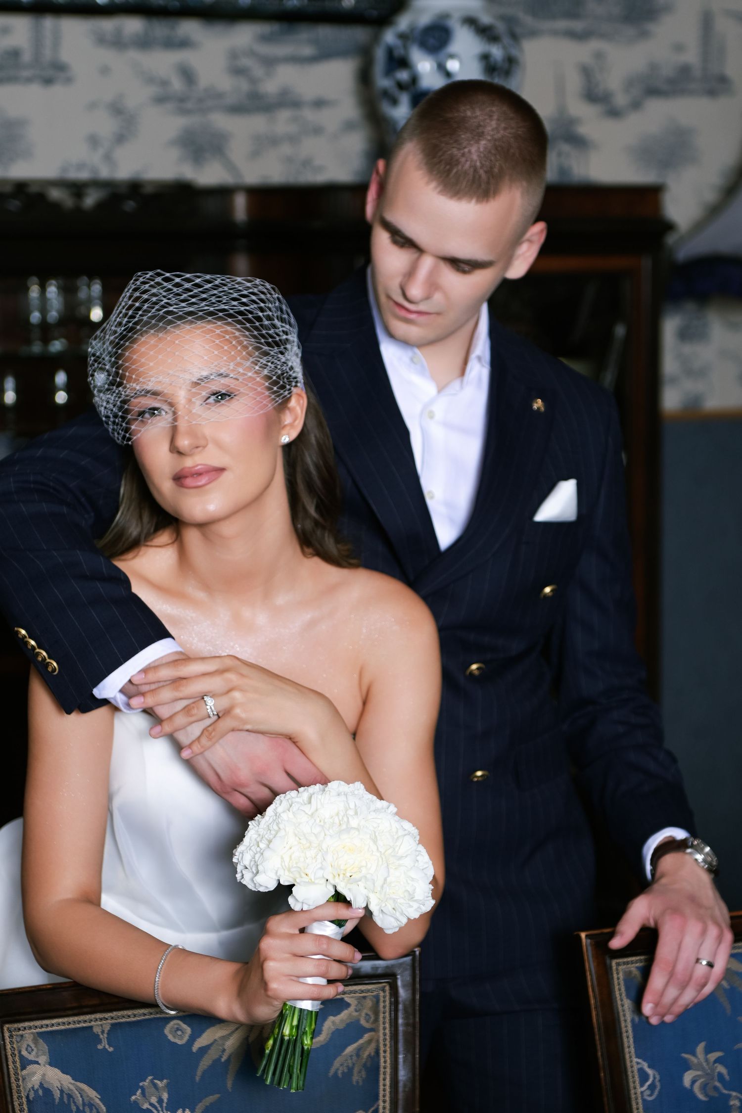 Elegant bride in white gown and birdcage veil holds white bouquet as groom in navy suit embraces her from behind.