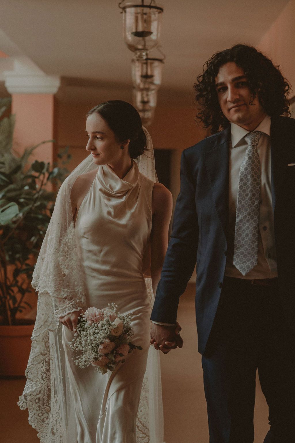 Bride in satin halter gown with lace veil holds bouquet while groom in navy suit holds her hand indoors.