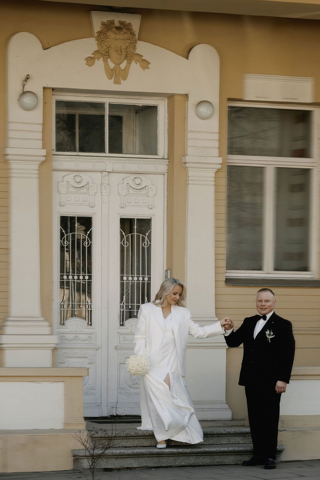 Bride in white pantsuit and groom in black tuxedo hold hands on steps of elegant yellow building.