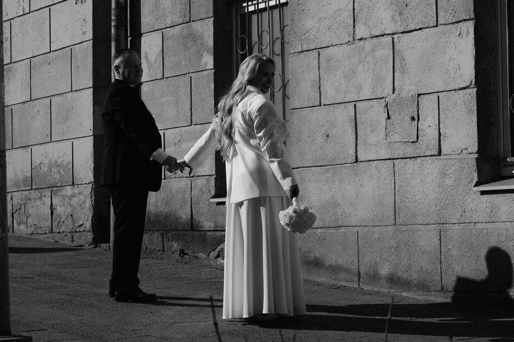 Black and white photo of a bride in white gown holding flowers, standing with a man in a suit near a stone wall.