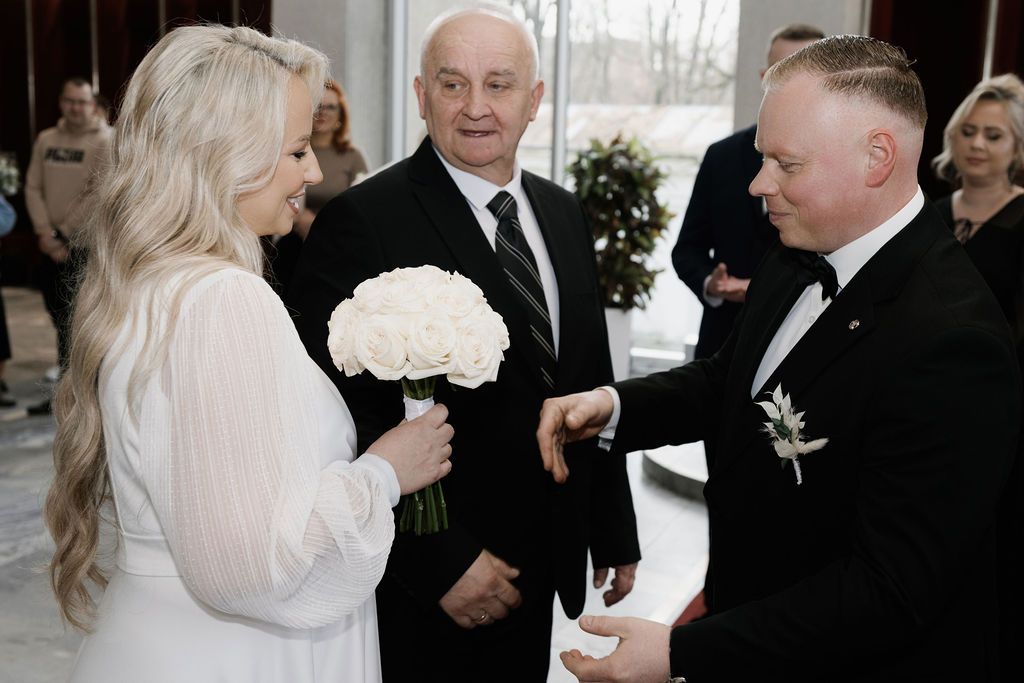 Bride in white dress holding bouquet exchanges vows with groom in tuxedo during elegant indoor wedding ceremony.