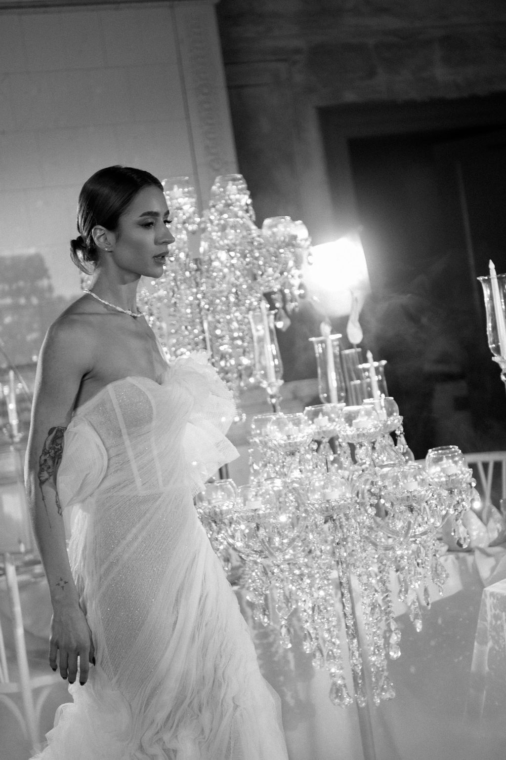 Black and white photo of a bride in a strapless gown standing beside crystal candelabras at an elegant wedding reception.