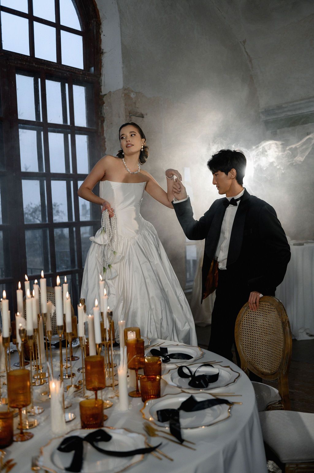 Bride in white gown stands on chair as groom in tuxedo raises glass beside candlelit wedding table.