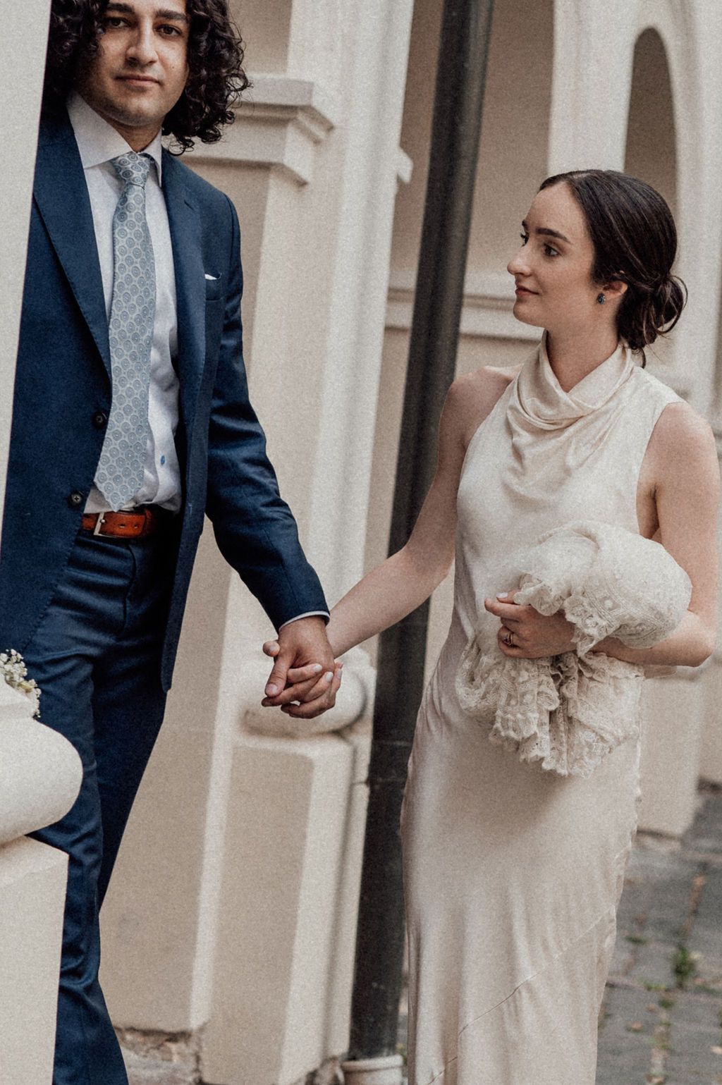 Wedding couple holding hands outdoors, groom in navy suit and bride in elegant ivory sleeveless gown with bouquet.
