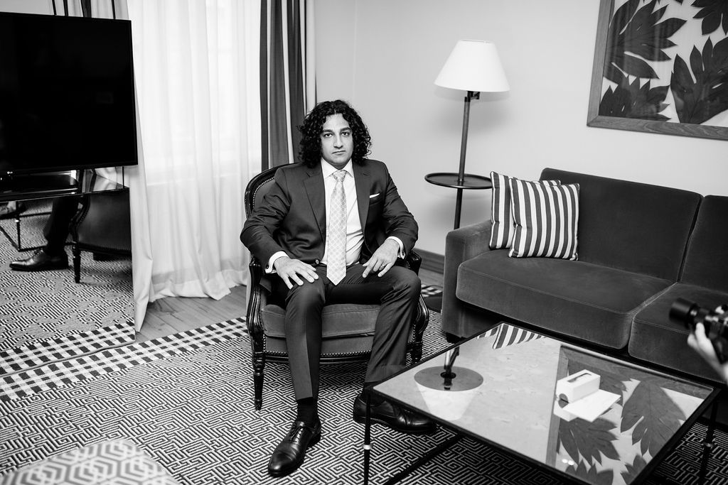 Stylish man in a suit sitting in a hotel room chair in a black and white portrait photo.