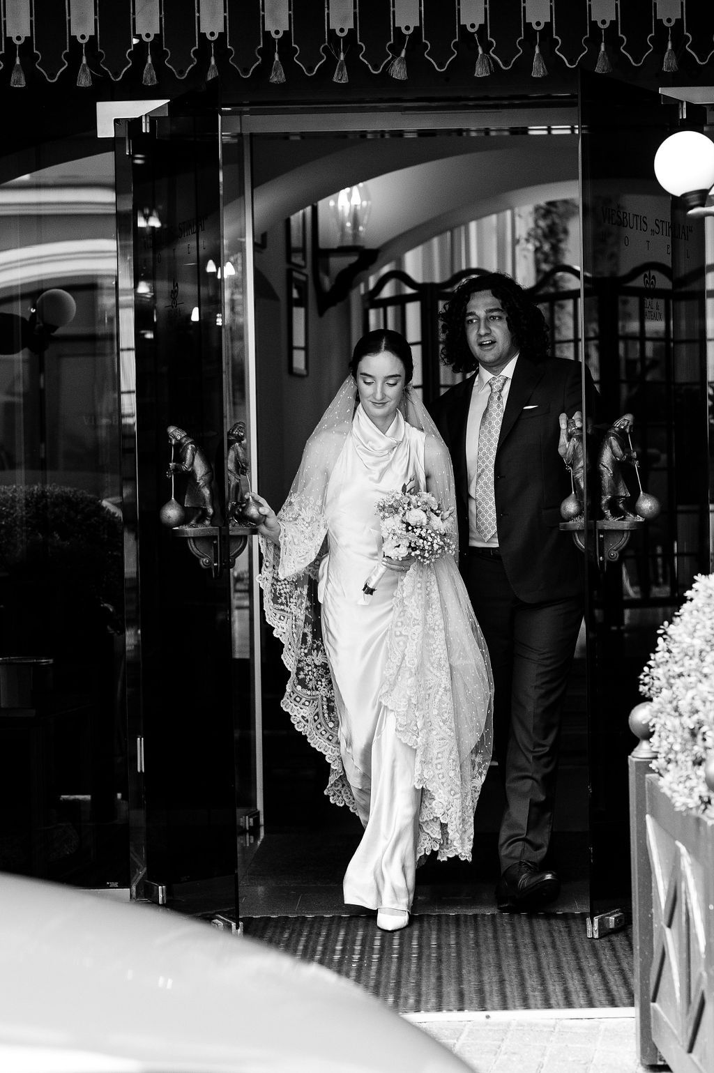 Bride in lace veil and groom in suit exiting a grand venue in a classic black and white wedding photo.
