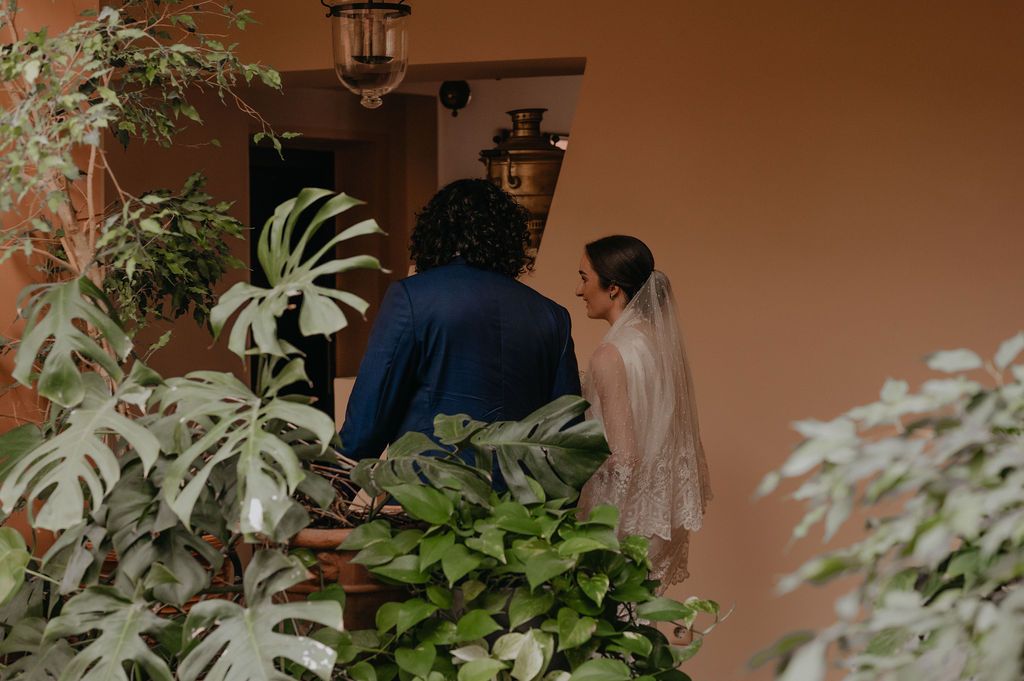 Bride and groom share an intimate moment at a terracotta villa entrance surrounded by lush monstera plants.