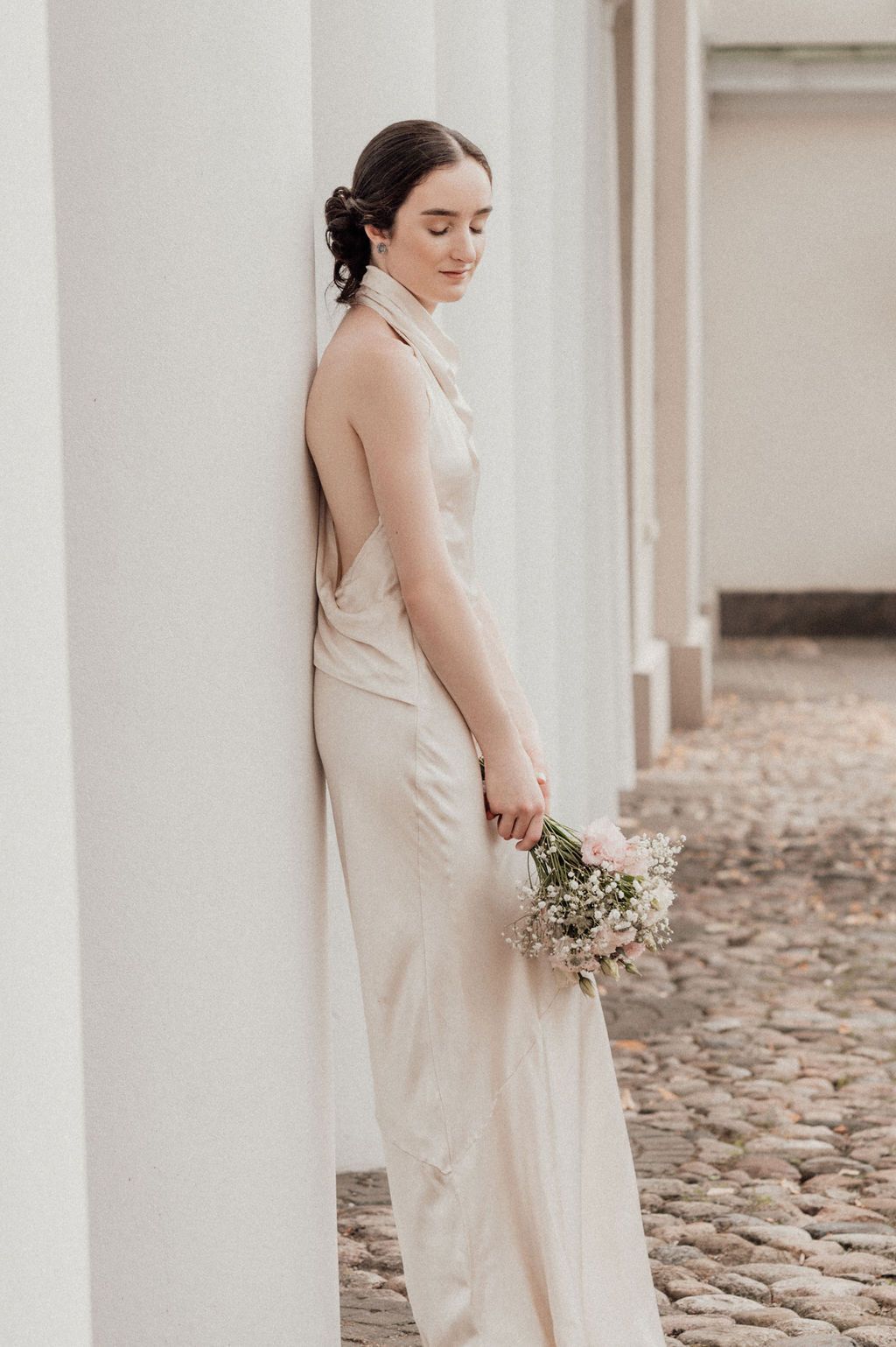Bride in backless halter satin gown holds bouquet leaning against white column on cobblestone path.