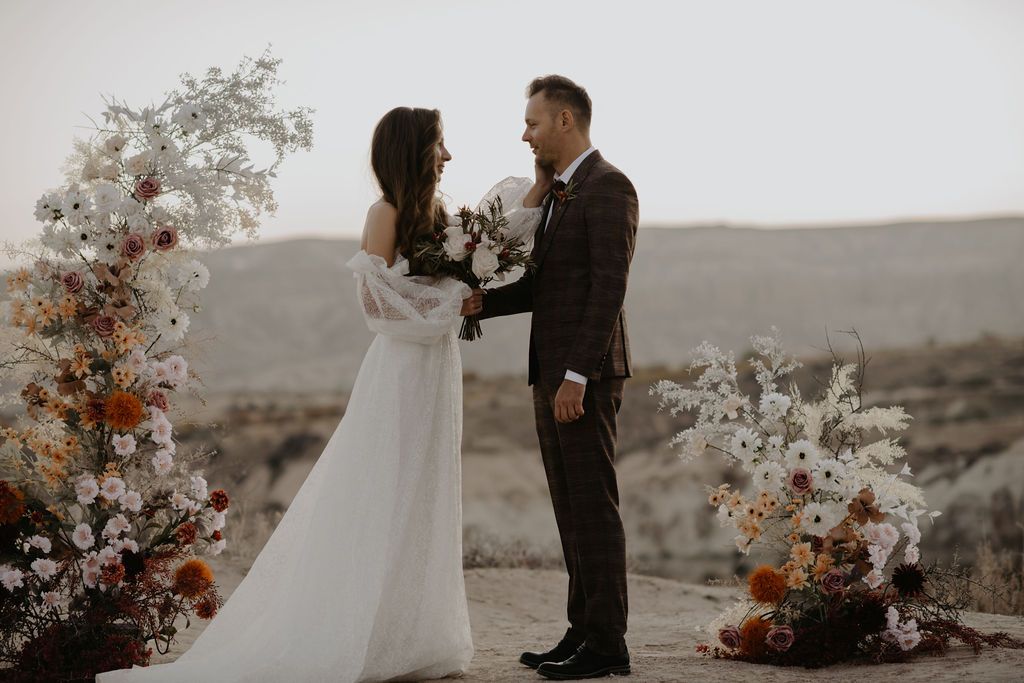 Bride and groom exchange vows outdoors in desert landscape, surrounded by elegant floral arrangements in white and terracotta hues.