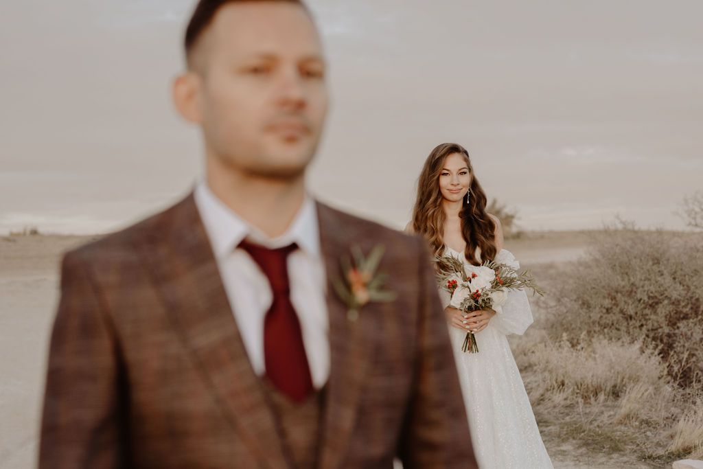 Groom in plaid suit with red tie stands in foreground as smiling bride holds bouquet in desert landscape.